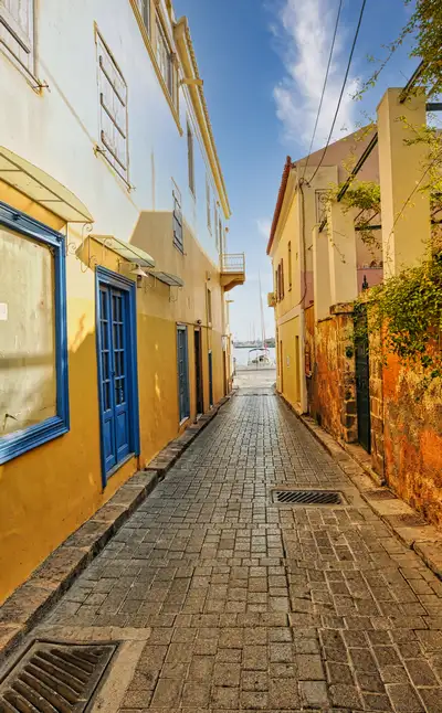 Narrow cobblestone alley in Aegina Town lined with colorful yellow and ochre buildings leading toward the harbor