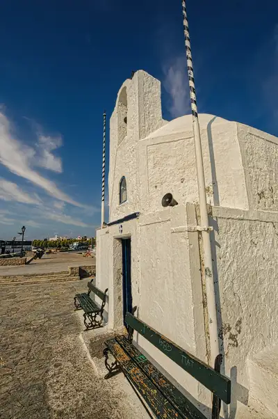 Small whitewashed chapel on the waterfront of Aegina Town with wooden benches outside and a deep blue sky above