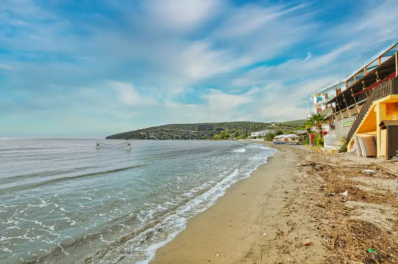 Sandy shoreline of Agia Marina beach in Aegina stretching along the coast with tavernas and green hills visible in the background