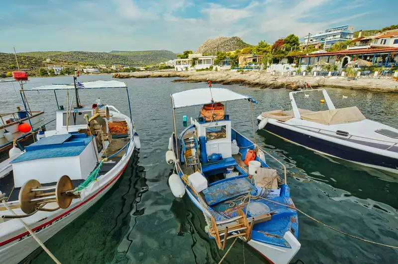 Small fishing boats moored in the sheltered harbor of Agia Marina village in Aegina with waterfront houses and green hills behind