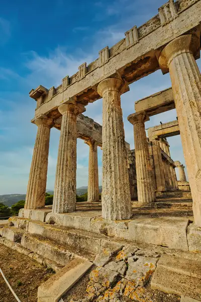 Close view of the Doric columns of the Temple of Aphaia in Aegina rising from ancient stone steps with the green hillside visible behind