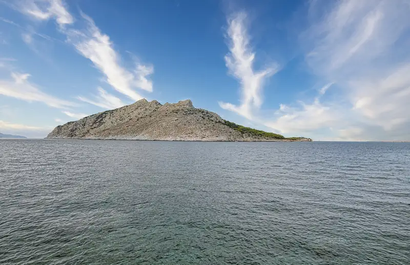 Uninhabited rocky island of Moni rising from the calm deep blue waters of the Saronic Gulf near Aegina, seen from across the sea