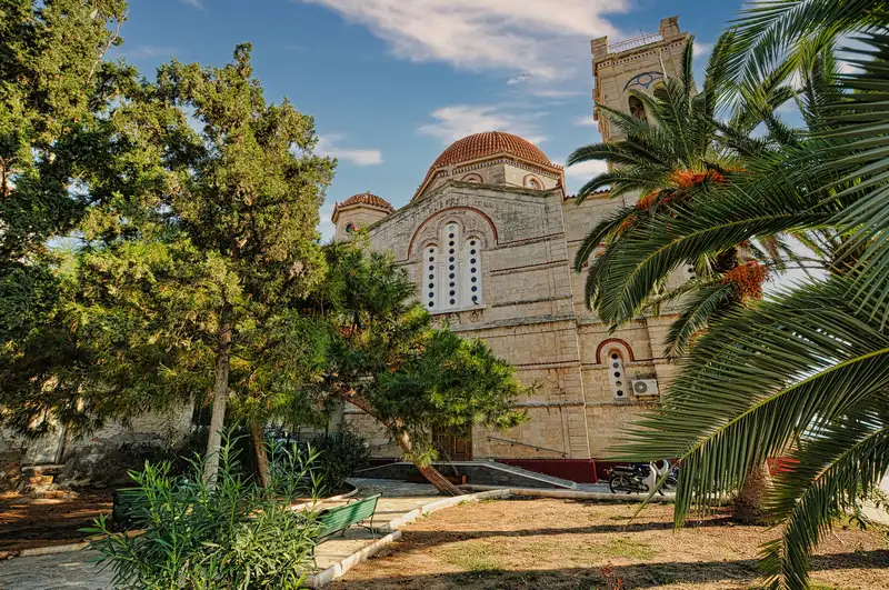Byzantine stone church with a terracotta dome in Aegina Town surrounded by tall pine trees and palm trees in a quiet garden setting