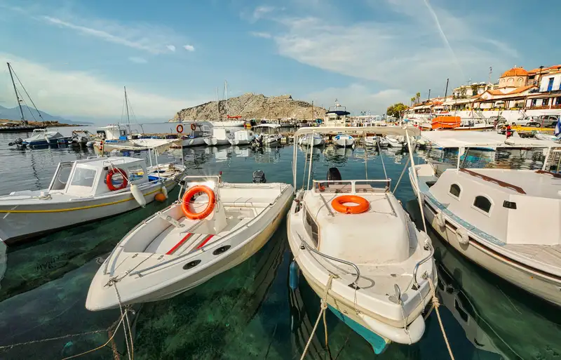 Small boats and motorboats moored in the calm harbor of Perdika port in Aegina with colorful waterfront buildings and a rocky hill in the background