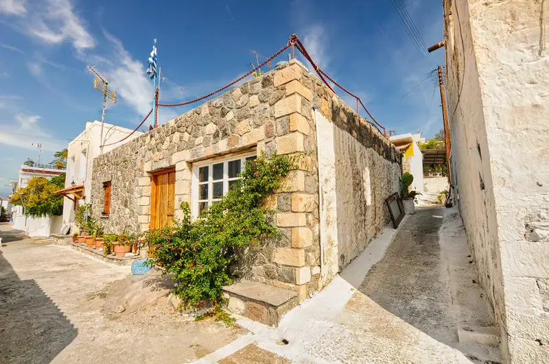 Traditional stone house with wooden door and flowering plants on a narrow whitewashed alley in Perdika village Aegina with a Greek flag above