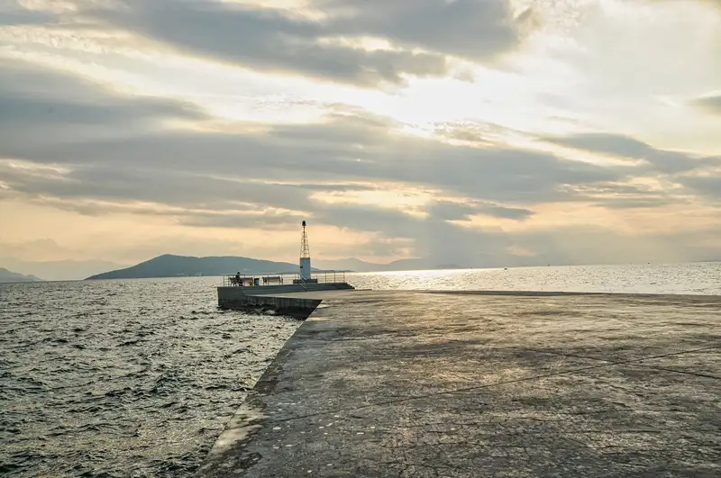 Long stone pier extending into the Saronic Gulf at sunset in Aegina with a solitary figure sitting at the end near a small lighthouse and an island silhouette in the distance