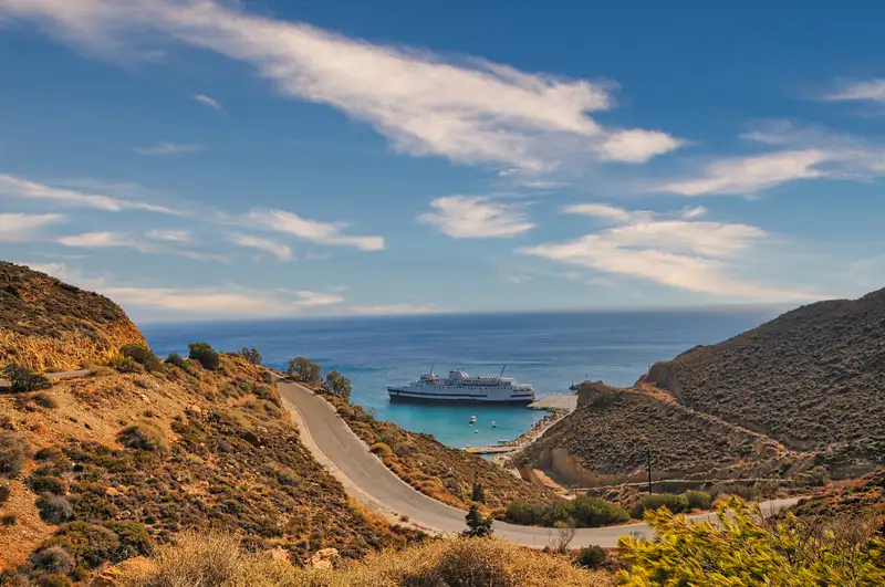 Winding coastal road leading toward Anafi port with a ferry docked in the bay below