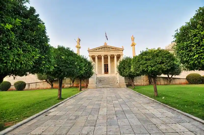 A tree-lined stone path leading to the entrance of the Academy of Athens, with orange trees on both sides