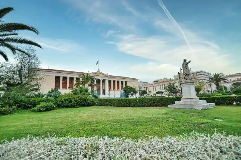 Wide view of the University of Athens main building from across the garden, with palm trees and a statue of a scholar in the foreground