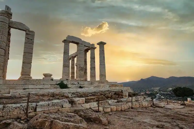 The ancient columns of the Temple of Poseidon at Cape Sounion standing on rocky ground with the sky glowing behind them