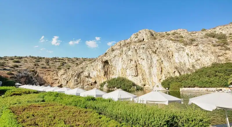 Lake Vouliagmeni near Athens with thermal waters, white umbrellas, lush green gardens, and dramatic limestone cliffs rising above