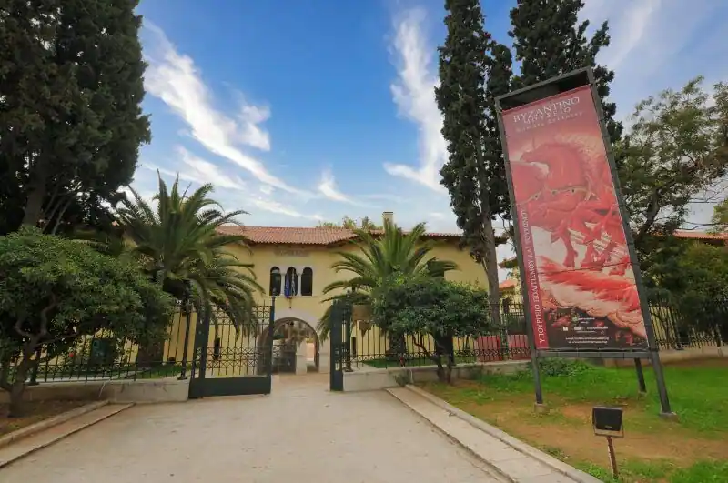 The main entrance of the Byzantine and Christian Museum in Athens with iron gates, palm trees, cypress trees, and Villa Ilissia visible behind the arched walkway