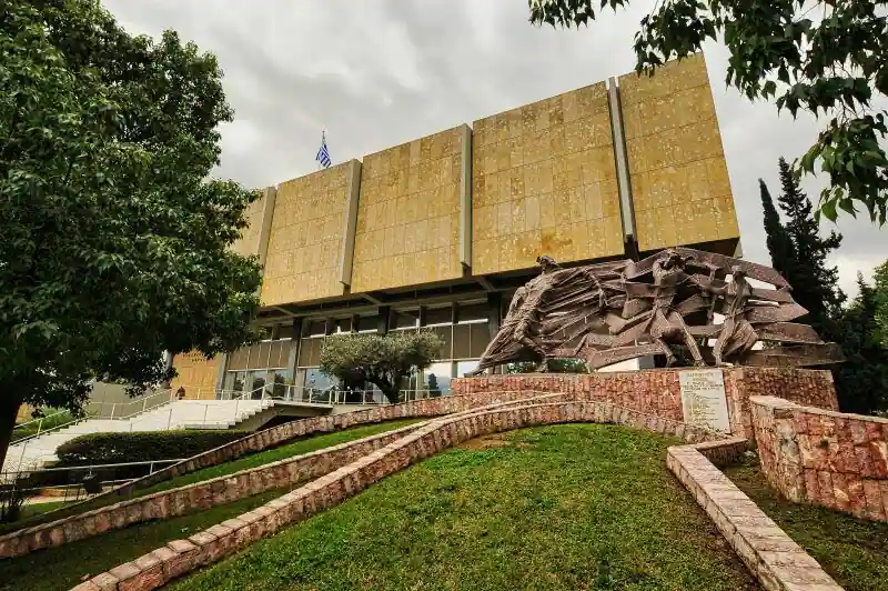 Exterior view of the War Museum Athens showing the Bauhaus style building with a bronze war memorial sculpture and green garden in front
