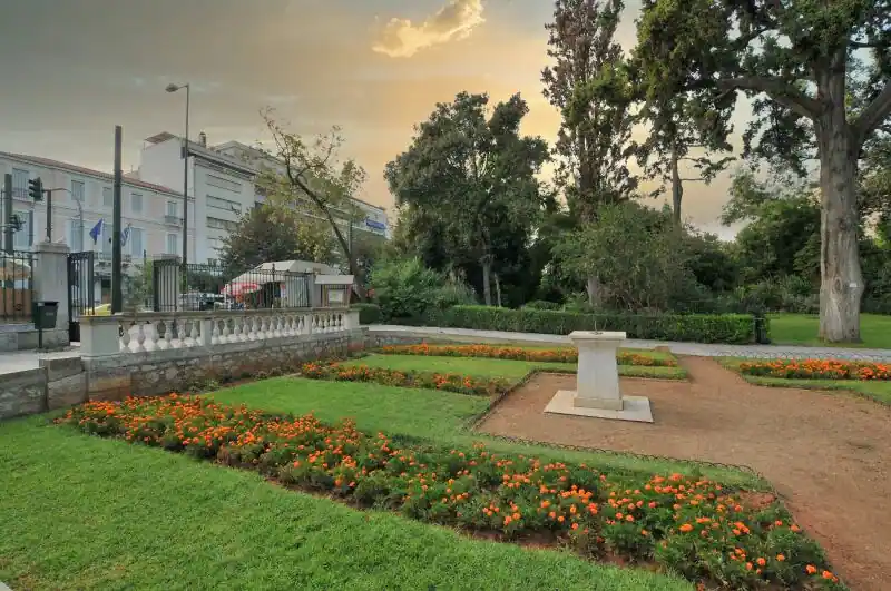 Entrance of the National Garden in Athens with orange flowers, green lawn, and tall trees at sunset