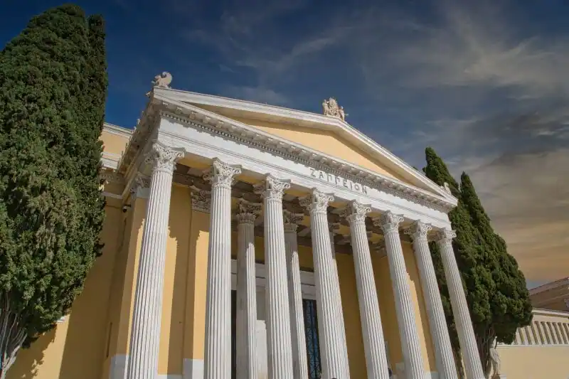 Neoclassical facade of the Zappeion Hall in Athens with tall white columns and cypress trees under a dramatic sky