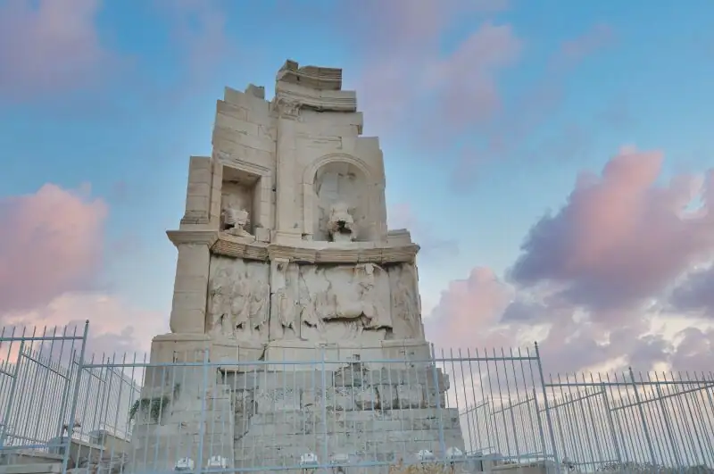 The front facade of the Philopappos Monument in Athens showing the carved frieze and statue niches under a pink and blue sky