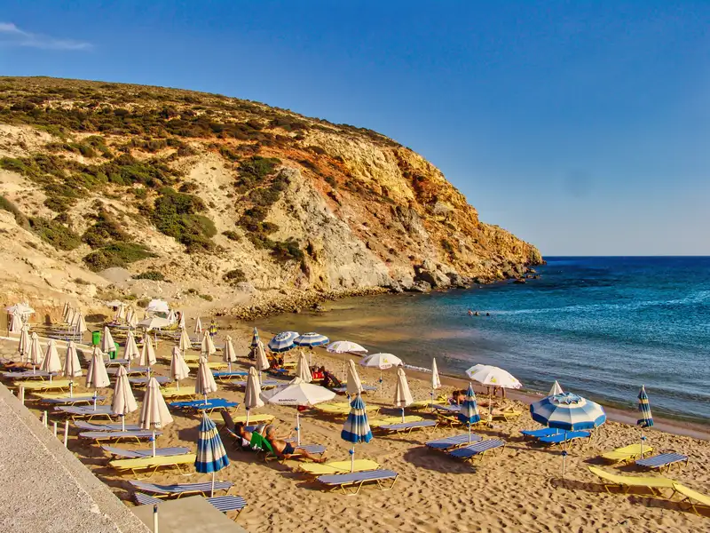 A sandy beach with sun loungers and striped umbrellas beneath an ochre cliff on Milos island