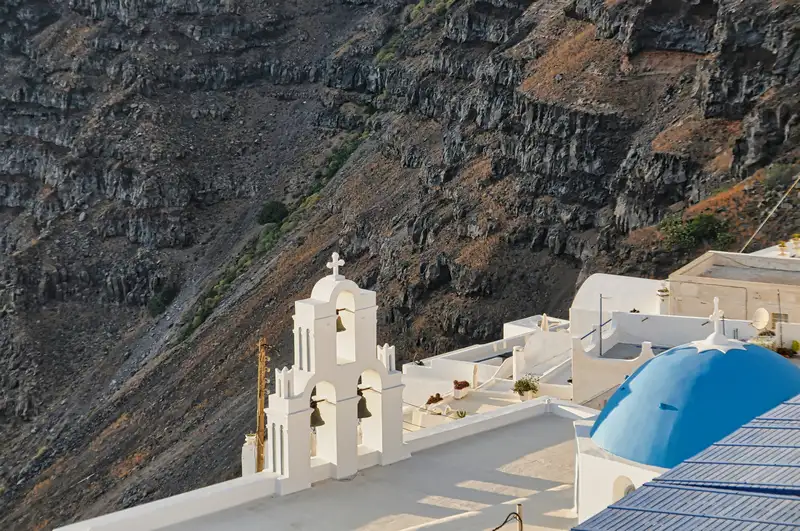 A whitewashed church with three bells below the dark cliffs of Santorini