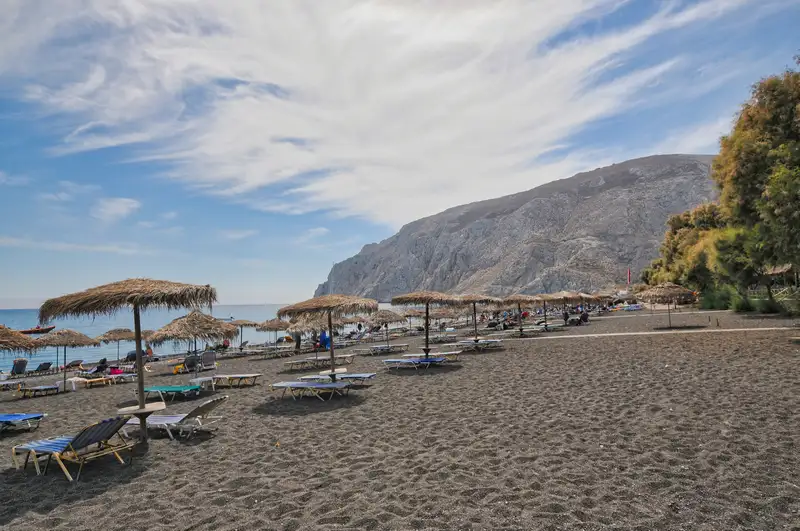 Black sand beach of Kamari in Santorini with thatched umbrellas below Mesa Vouno