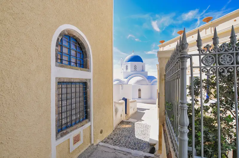 A blue-domed church in Pyrgos, Santorini, seen down a stone lane between a yellow wall and an iron gate