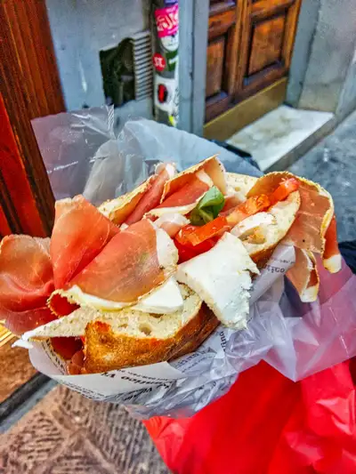 Hand holding a Tuscan schiacciata sandwich filled with prosciutto, cheese, and tomato in Florence