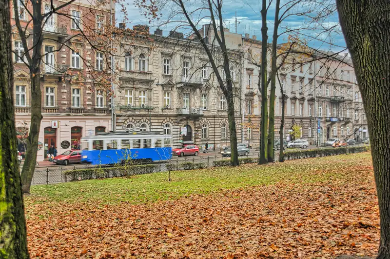 Blue tram passing old tenement houses beside a leaf-covered park in Krakow